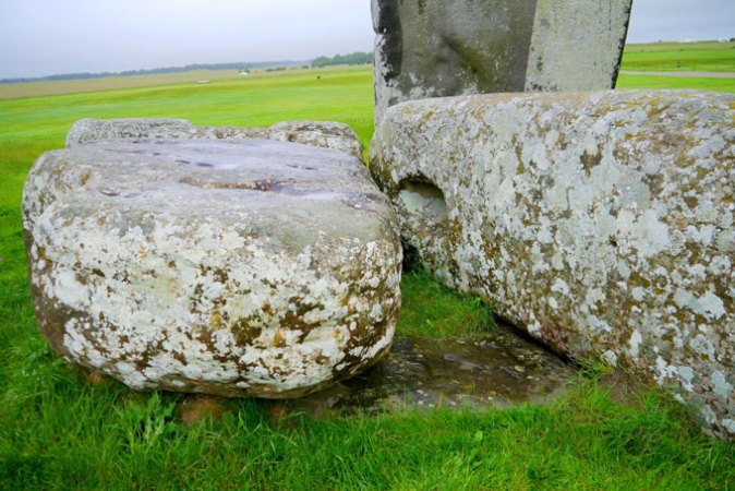 In this close-up photo, two mottled stones from Stonehenge rest on top of a darker brown stone embedded in the grass. That partially buried stone is the Altar Stone.